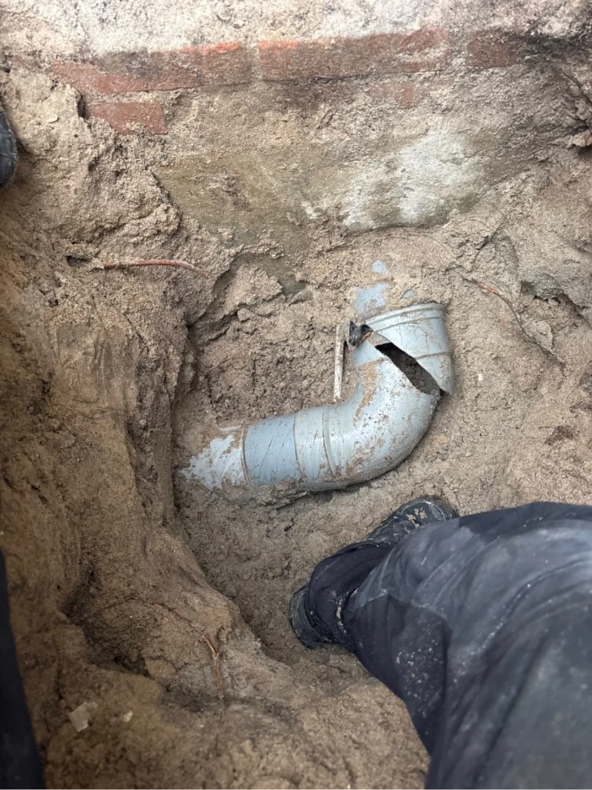 Opgraving in zandgrond toont ernstig gebroken PVC-afvoerbuis (bochtstuk) rioleringssysteem.