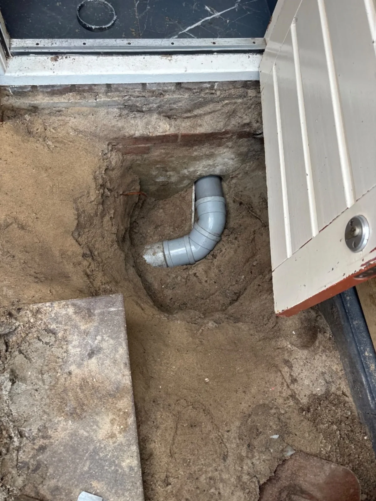 Uitgegraven gat onder vloer toont grijze PVC afvoerbuis met 90-graden bocht, zand en bakstenen.